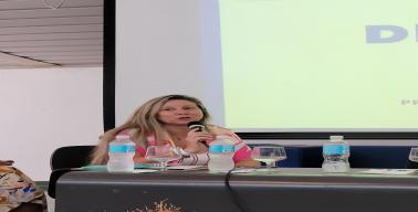 Descrição da imagem: Foto da professora Micheline sentada à frente da bancada. Ela segura o microfone e gesticula com a boca. A professora Micheline é branca, tem cabelos lisos e loiros e está com uma blusa florida.